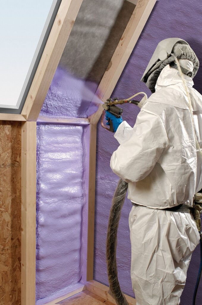 A person wearing a protective suit, goggles, and a face mask while carrying a foam sprayer to spray foam insulation for a steel building